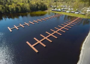 An aerial view of a large floating dock system made with AlumaDeck decking, featuring a long central walkway with multiple perpendicular docking sections extending over a calm lake, surrounded by greenery and waterfront properties.