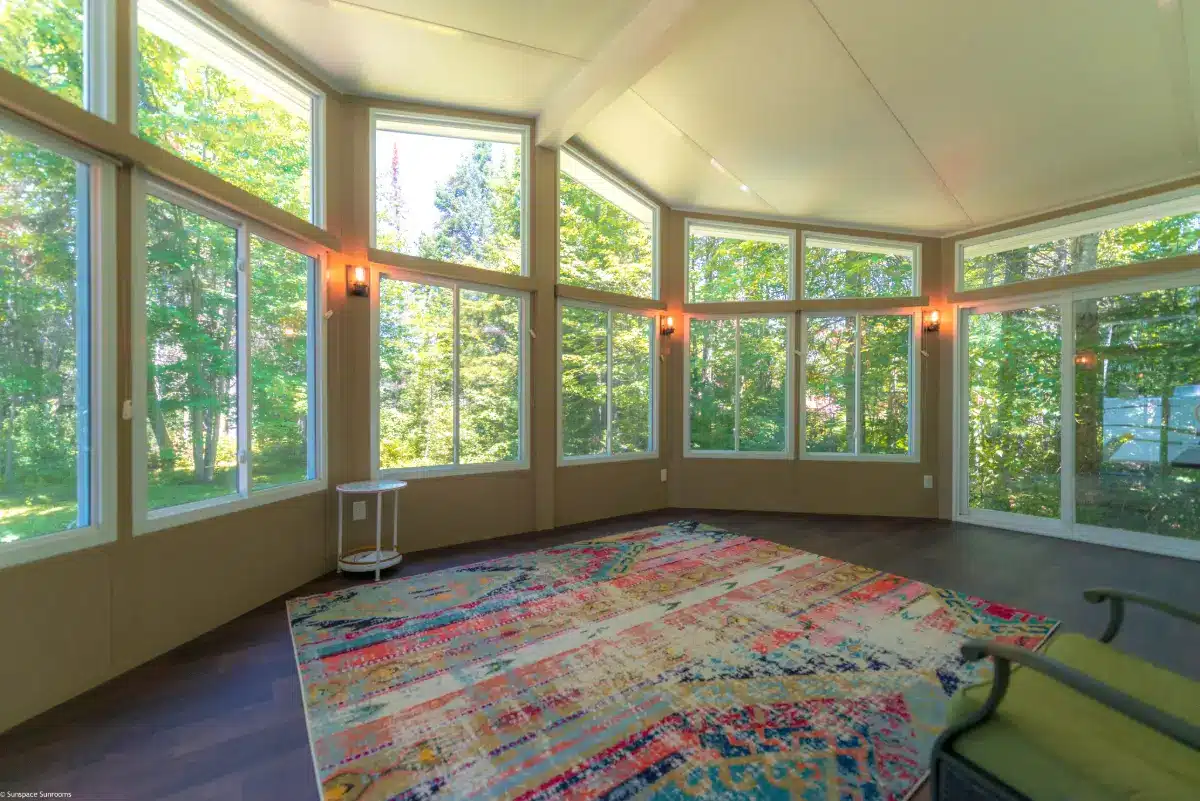 A spacious sunroom with large glass windows, a vaulted ceiling, and warm lighting, offering stunning views of lush greenery outside.