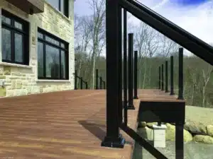 An elevated modern deck with a wood-look surface, black metal railing posts, and a strong support structure, attached to a stone-clad house with a scenic forest view.