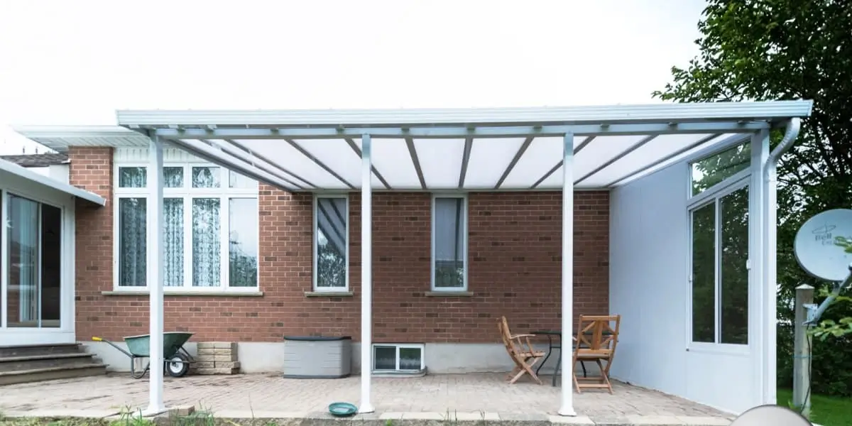 A modern acrylic patio cover with a white aluminum frame attached to a brick house, providing shade and weather protection while allowing natural light to pass through.