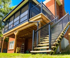 Elevated Sunspace WeatherMaster™ Model 200 sunroom with black aluminum framing, vertical sliding windows, and a wooden deck with a staircase.