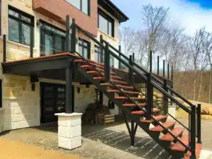 A modern outdoor decking system featuring AlumaDeck aluminum decking with glass railings, black metal framing, and wooden steps, attached to a contemporary home with stone and wood siding.
