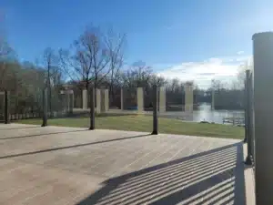 Topless glass railing on an outdoor deck, featuring black metal posts with a scenic view of green grass, trees, and a flowing river in the background